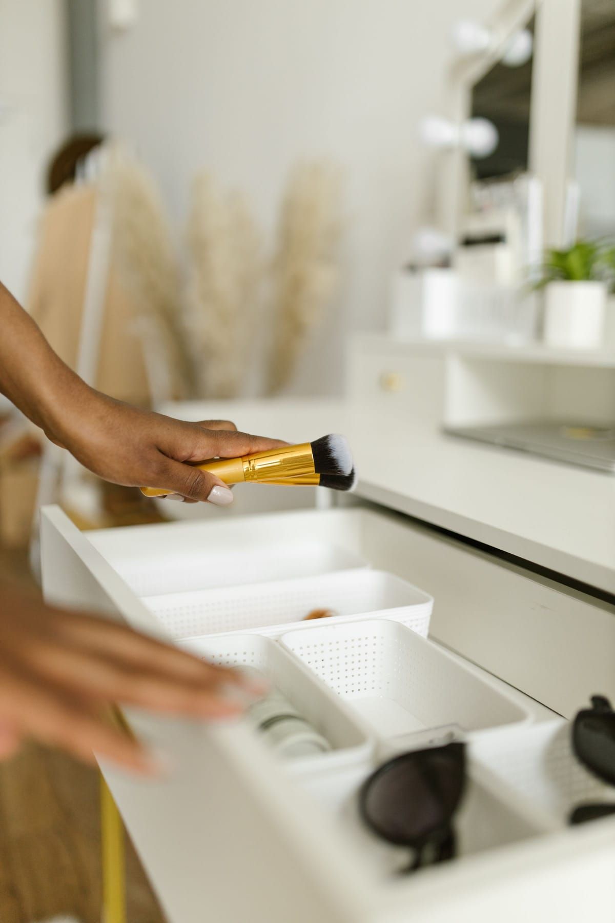 A hand holding a makeup brush over an open white vanity drawer lined with small organizer bins