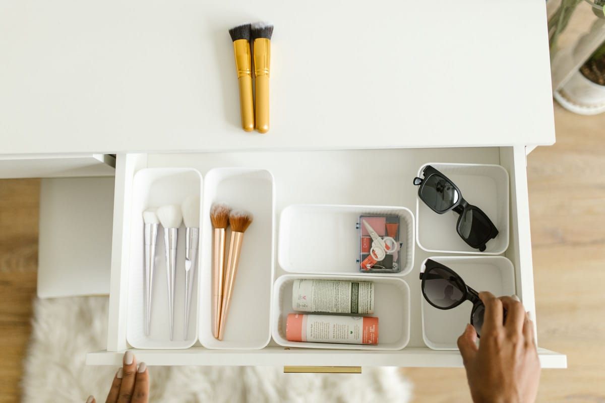 An open vanity drawer with white organizer trays holding makeup brushes, skincare tubes and sunglasses