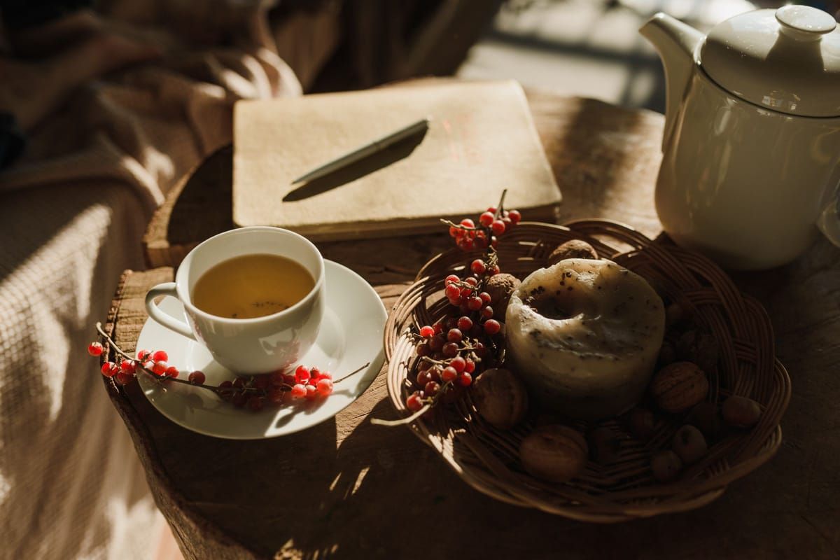 A cup of tea, a notebook and pen, and a white teapot on a wooden table in warm afternoon light