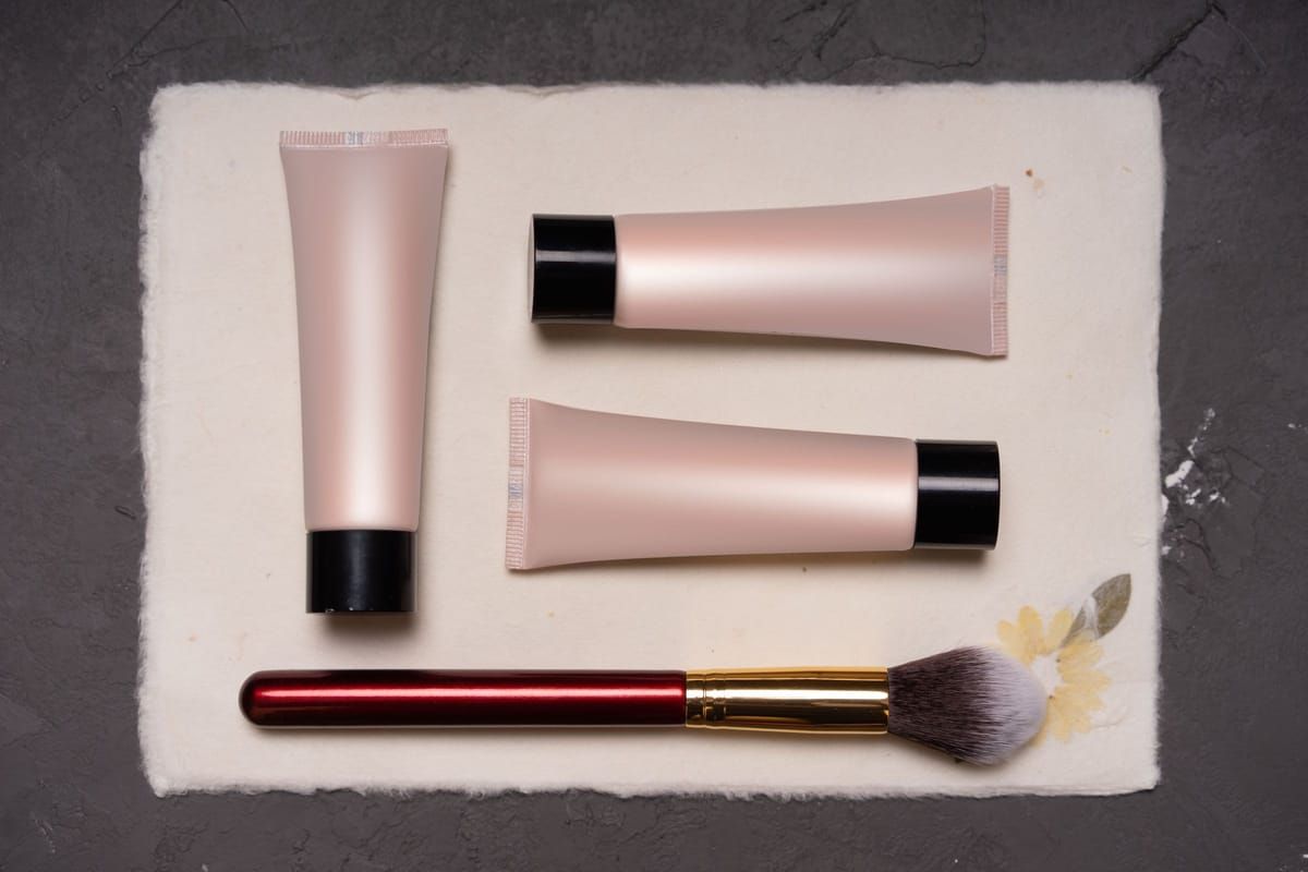 Three soft pink makeup tubes and a fluffy brush laid out on cream paper
